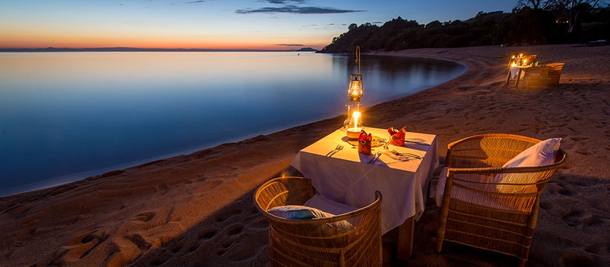 Romantic private dining setup on the sandy shore of Lake Malawi illuminated by lanterns at dusk.