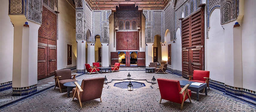 Grand courtyard lounge at Dar Bensouda featuring ornate Moroccan tilework, carved wood, and red accent chairs.