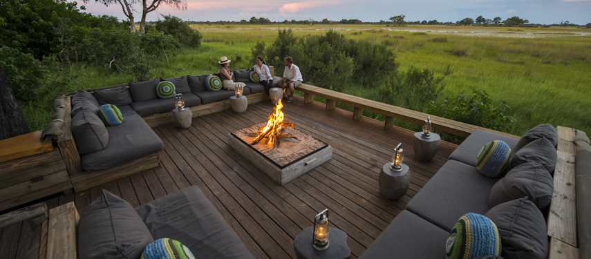 Guests gathered around a campfire at sunset on a timber verandah at Vumbura Plains Camp, Botswana
