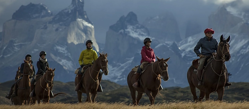 A group of riders, led by a local gaucho, traverse open grasslands beneath brooding Patagonian mountains.