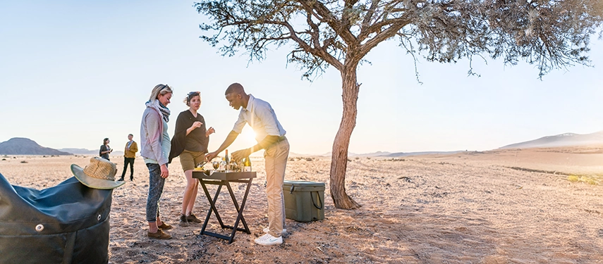 Morning light illuminating a breakfast setup under a solitary tree at Camp Sossus as guests gather in the Namib Desert.