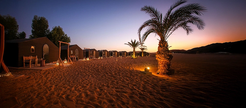 Row of luxury desert tents illuminated at dusk with palm trees in the sand at Riad Madu Camp.