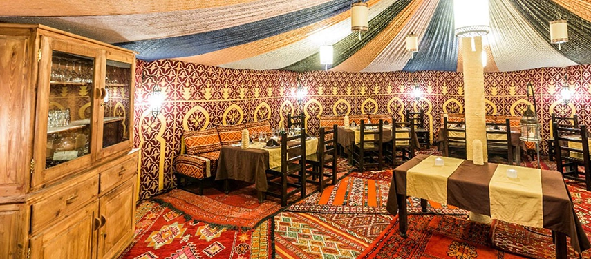 Colourful Moroccan dining tent with patterned walls, low lighting, and traditional carpets at Riad Madu Luxury Camp.