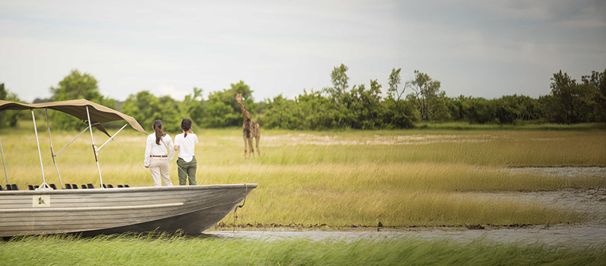 Two guests on a boat safari along the Chobe River observing a giraffe in the distance across open grassy floodplains.