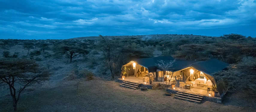 An aerial view of two illuminated canvas tents at Hemingways River Camp Mara glowing warmly against the darkening Mara North Conservancy landscape at dusk.