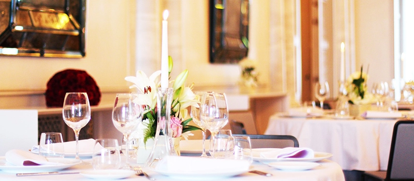 Elegant dining tables with candles and flowers set for a formal event in a softly lit restaurant.