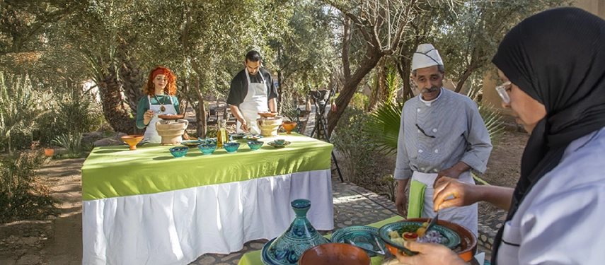 Outdoor Moroccan cooking class at Ksar El Kabbaba with chefs and guests preparing tagines beneath olive trees.