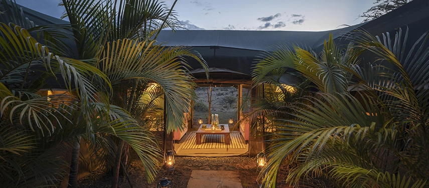 The lantern-lit entrance to the Family Tent at Hemingways River Camp Mara at dusk, framed by lush tropical palms and a glimpse of the Mara beyond.