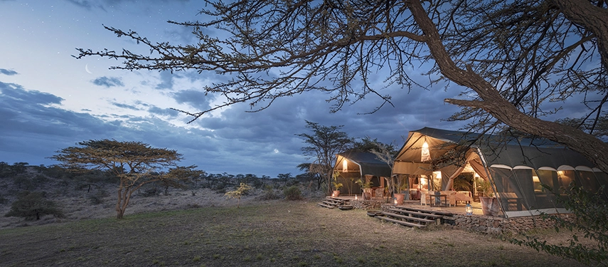 The Family Tent at Hemingways River Camp Mara illuminated at dusk beneath a crescent moon, its open veranda and warm interior light spilling into the surrounding Mara bush.