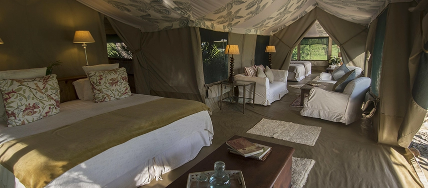 A generously proportioned Family Tent interior at Hemingways River Camp Mara, with twin beds, floral cushions, soft lamplight and a sitting area opening to the bush beyond.
