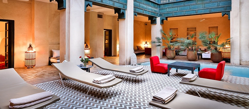 Courtyard poolside at Dar Bensouda with loungers, palm plants, and vibrant red chairs.