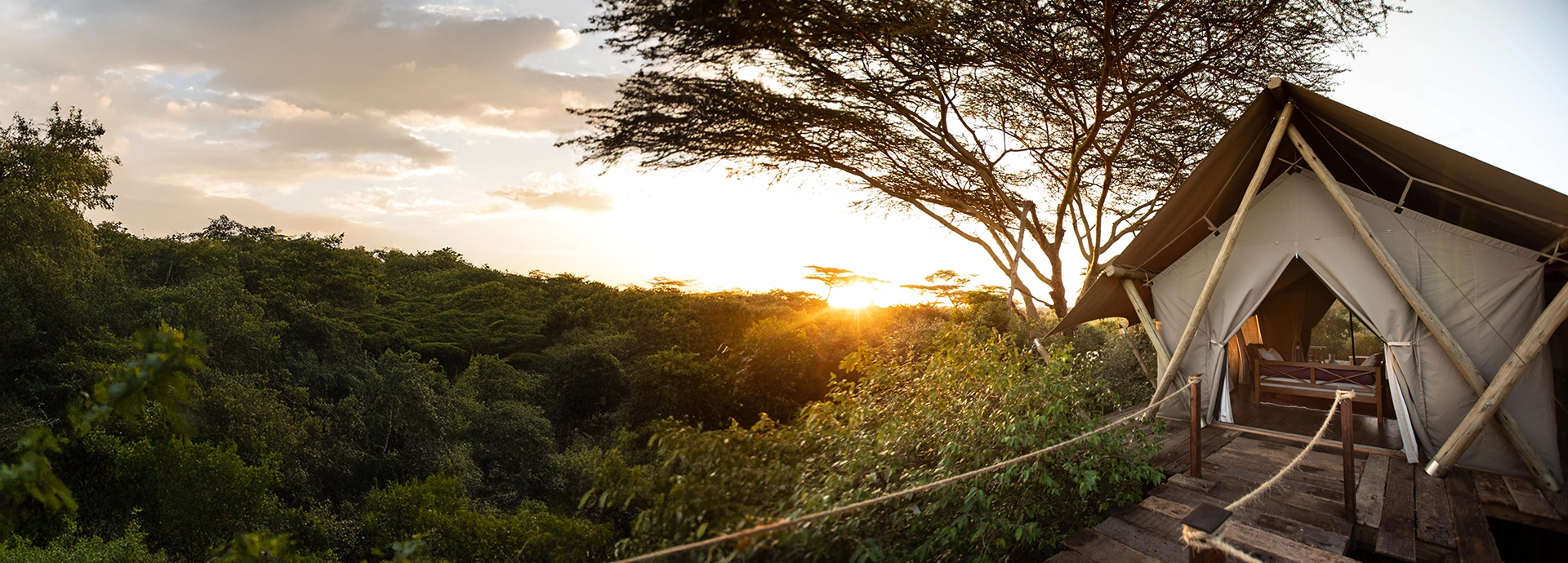 Exterior view of a canvas tent at Mara Nyika Camp in Kenya’s Maasai Mara, overlooking dense riverine forest as the sun sets across the savannah.