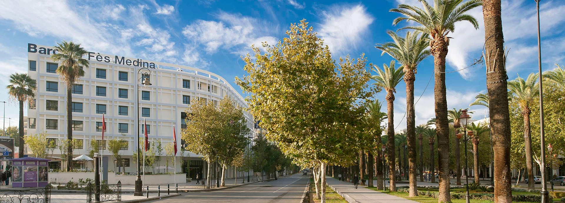 Wide boulevard lined with palm trees leading towards Barceló Fès Medina under a bright blue sky.