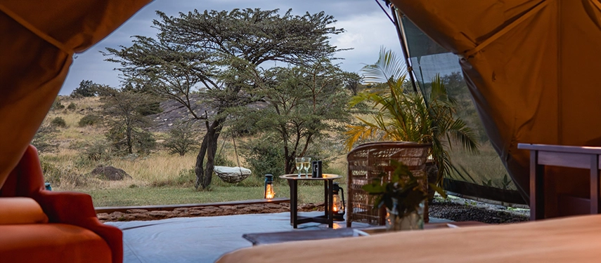 The view from inside a Family Tent at Hemingways River Camp Mara, looking out through open canvas walls to a hammock, lanterns and the rocky acacia-studded landscape at dusk.