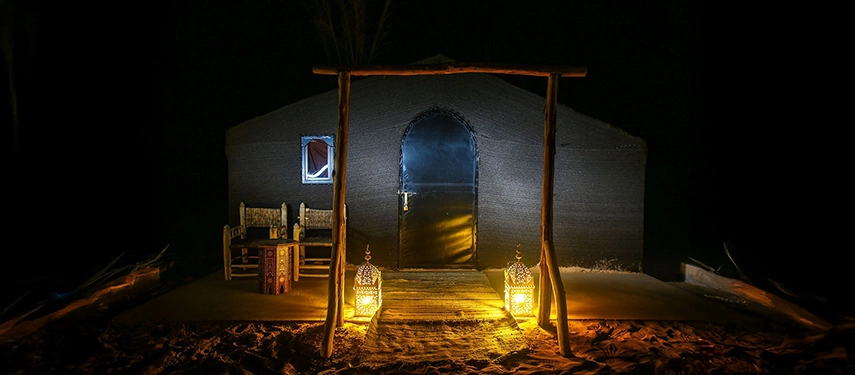 Luxury desert tent entrance lit by lanterns at night with wooden chairs on the sandy terrace.