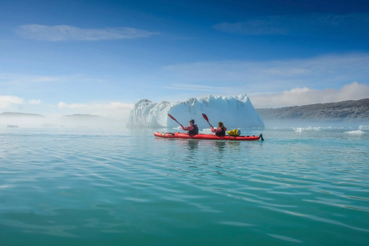 Two kayakers paddling past a towering iceberg under clear Arctic skies, capturing the serenity of polar exploration.