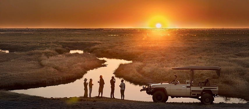 Safari group enjoys a sundowner by a remote river bend as the sun sets over the Linyanti wetlands.