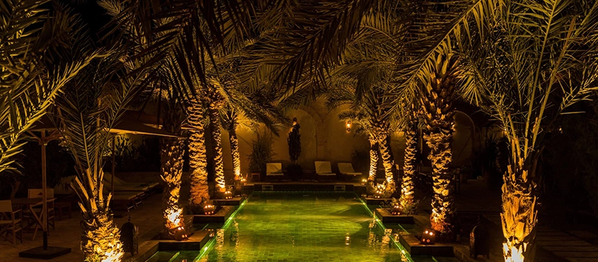 Illuminated courtyard pool at Dar Tozeur framed by tall palm trees and glowing lanterns at night.