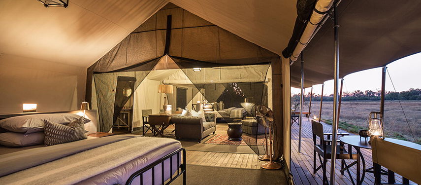 Elegant safari tent interior with a queen bed, canvas partitions, soft lighting, and views onto the bush from the wooden deck.