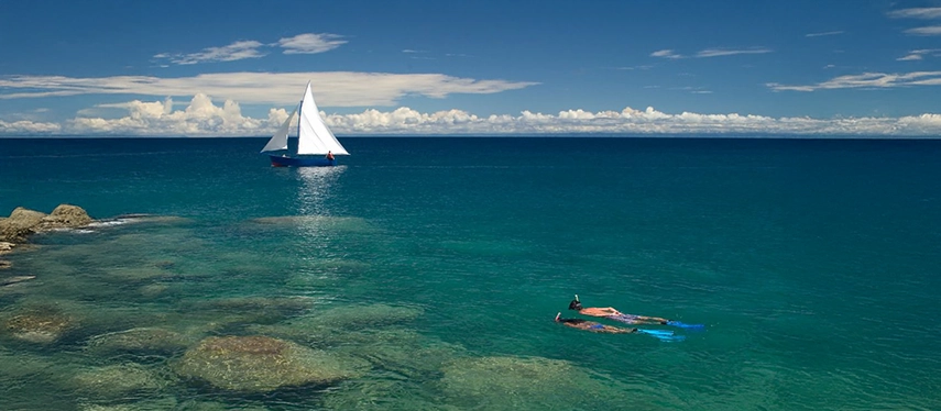 Snorkellers exploring crystal-clear waters of Lake Malawi with a white-sailed dhow in the distance.