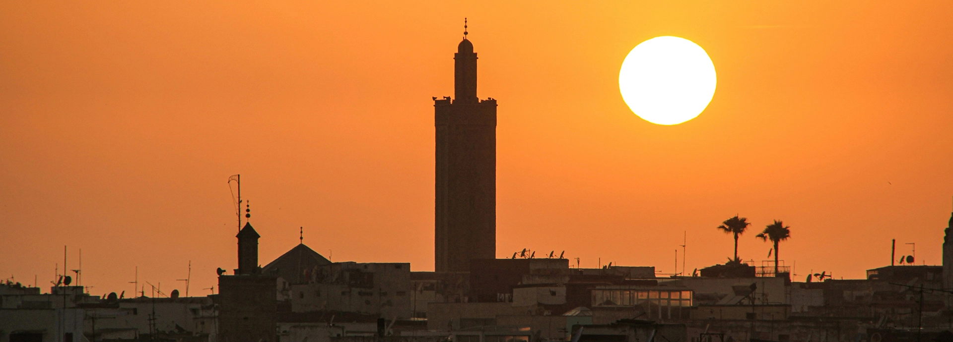 Golden sunset over Rabat with the silhouette of a mosque minaret and palm trees against an orange sky.