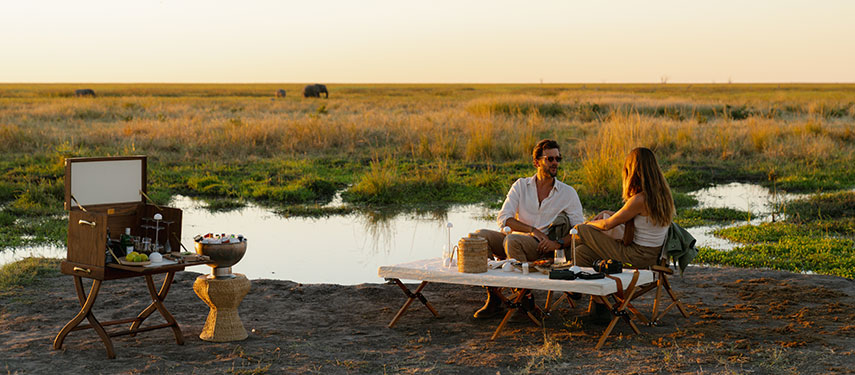 Couple enjoy sundowners next to a pool with elephant in the background on safari in Botswana