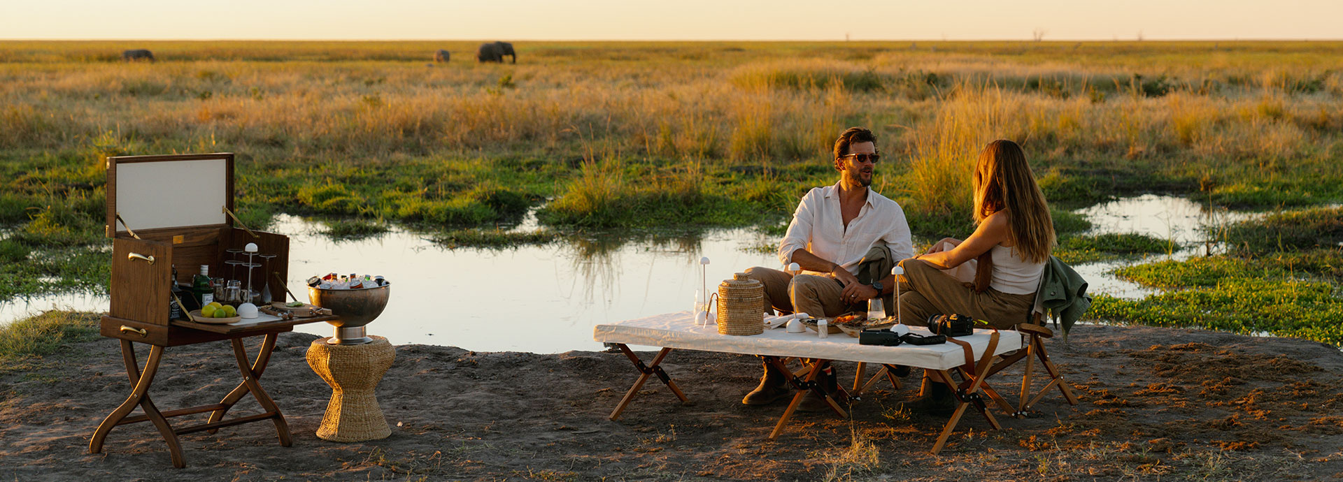 Couple enjoy sundowners next to a pool with elephant in the background on safari in Botswana