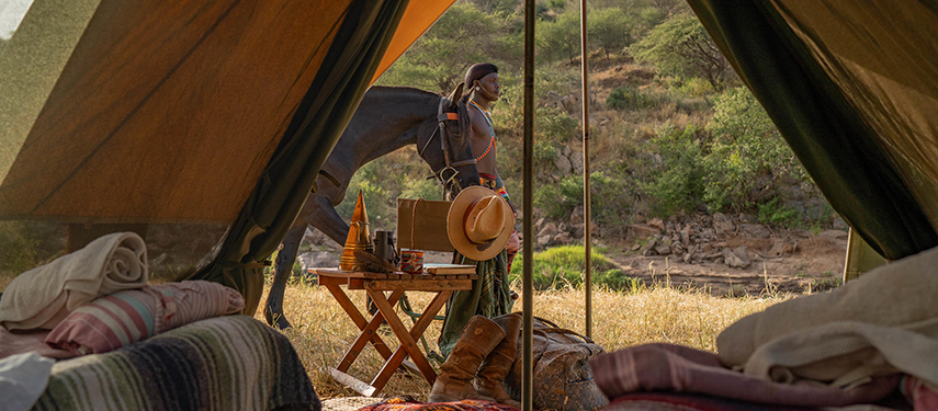 A Samburu warrior stands beside a saddled horse outside a tent, viewed from within a fly camp setup with rustic furnishings.