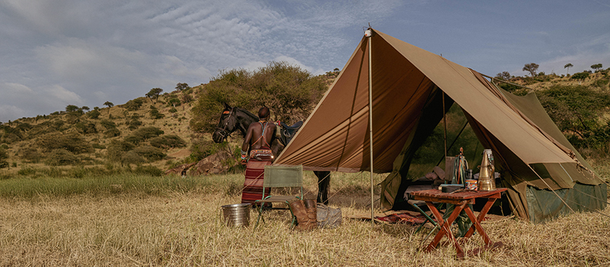 A traditional canvas tent pitched in a grassy clearing, with a Samburu guide and horse standing beside outdoor camping gear.