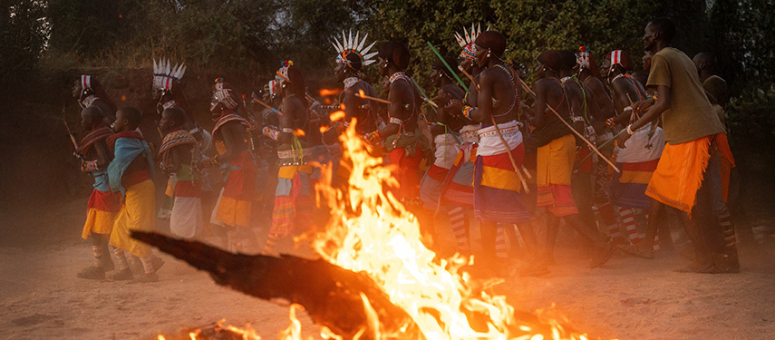 A vibrant traditional Samburu dance takes place around a fire, with men and women dressed in colourful beadwork and ceremonial attire.