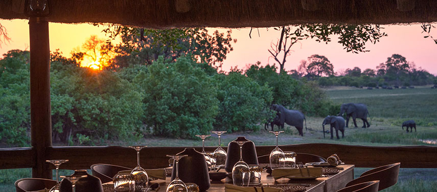 Dining area at Savuti luxury safari camp at sunset with elephants grazing under the background