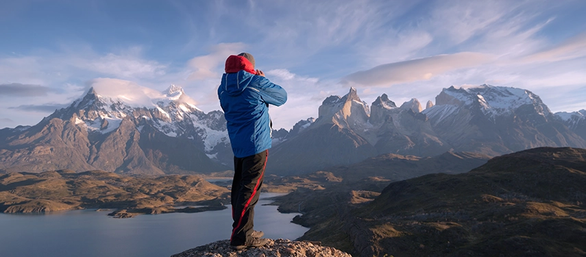 A traveller photographing the snow-capped Torres del Paine massif from a high Patagonian viewpoint at sunrise.