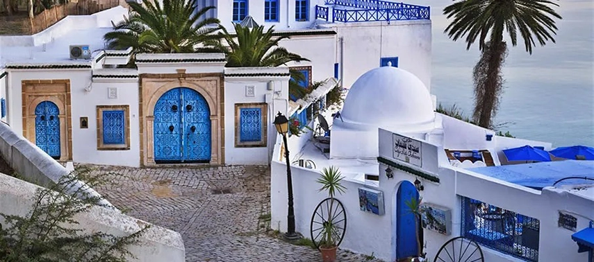 Traditional cobbled street with bright blue doors, whitewashed walls, and a domed rooftop overlooking the sea.