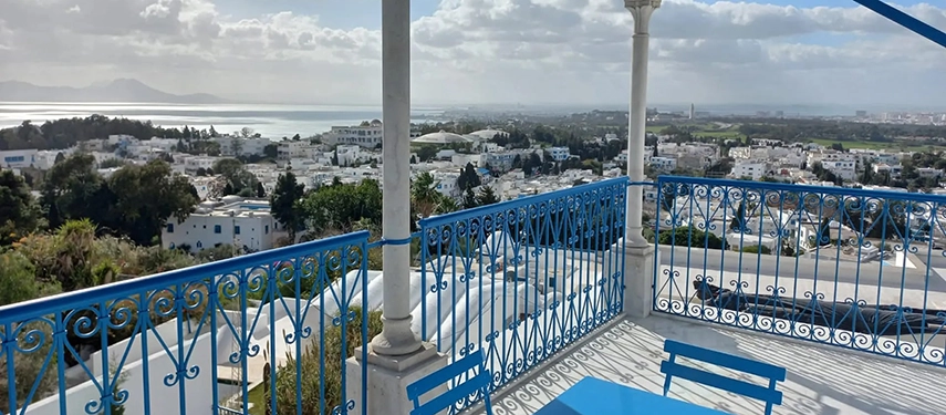 Balcony with blue wrought-iron railing and table set, overlooking sweeping views of rooftops and coastline.