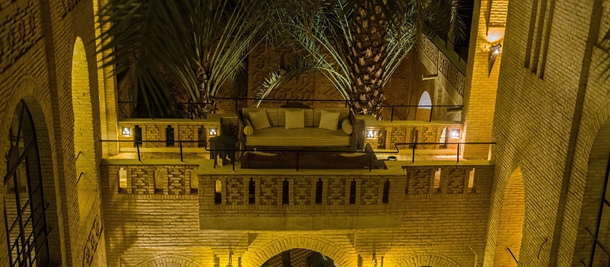 Romantic riad courtyard at night with a sofa lounge surrounded by palm trees and golden lighting.
