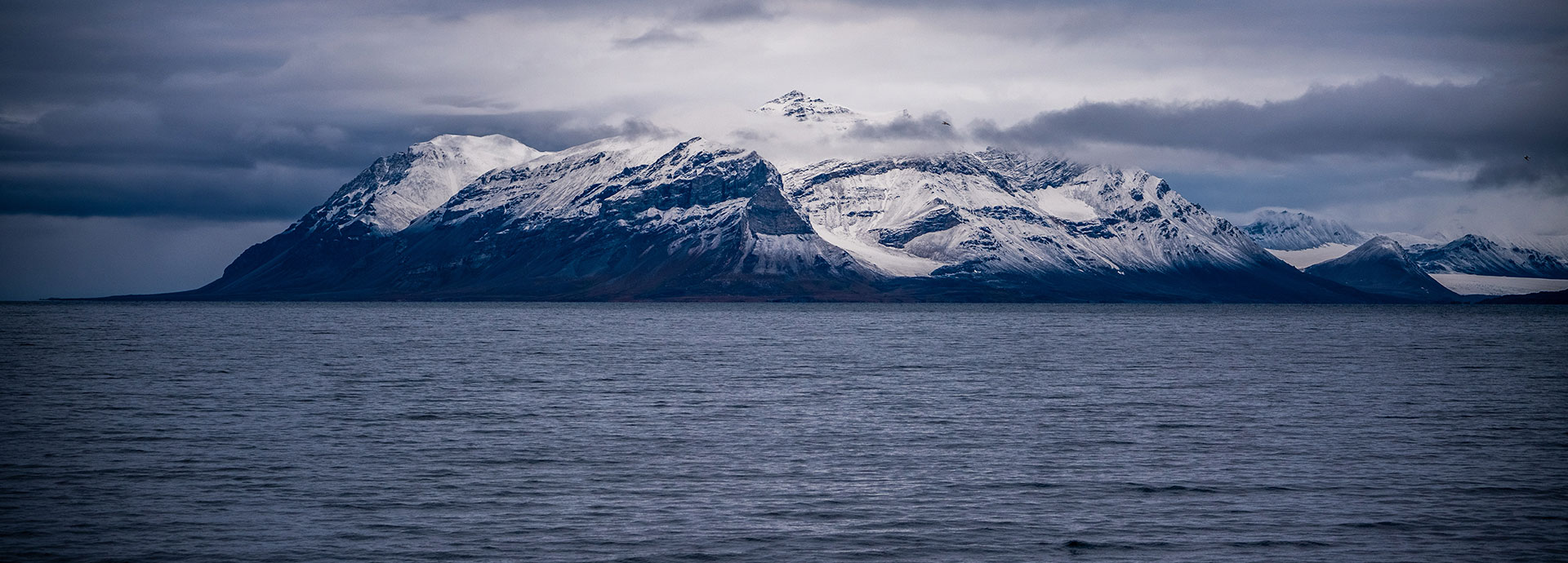 Snow-dusted mountain rising from the ocean under a moody Arctic sky, showcasing the remote wilderness explored on the Jewels of the Arctic expedition.