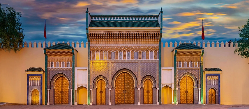 Royal Palace of Fès with golden gates, intricate mosaic arches, and green tiled roofs illuminated at dusk.