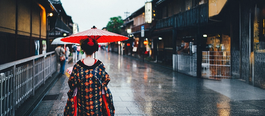 A woman in an ornate kimono walks through a historic Kyoto street in the rain, holding a red paper umbrella.