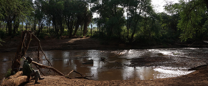 A guide and guest sit by a river on an African walking safari